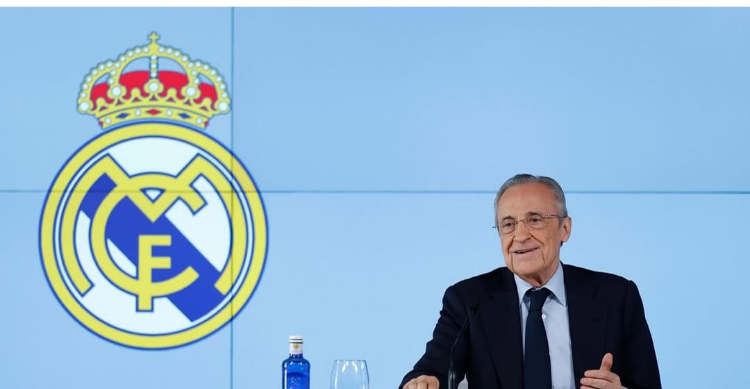 O presidente do Real Madrid de terno e gravata preta e camisa branca está à direita da foto. Ao seu lado, na parede o símbolo do Real Madrid.